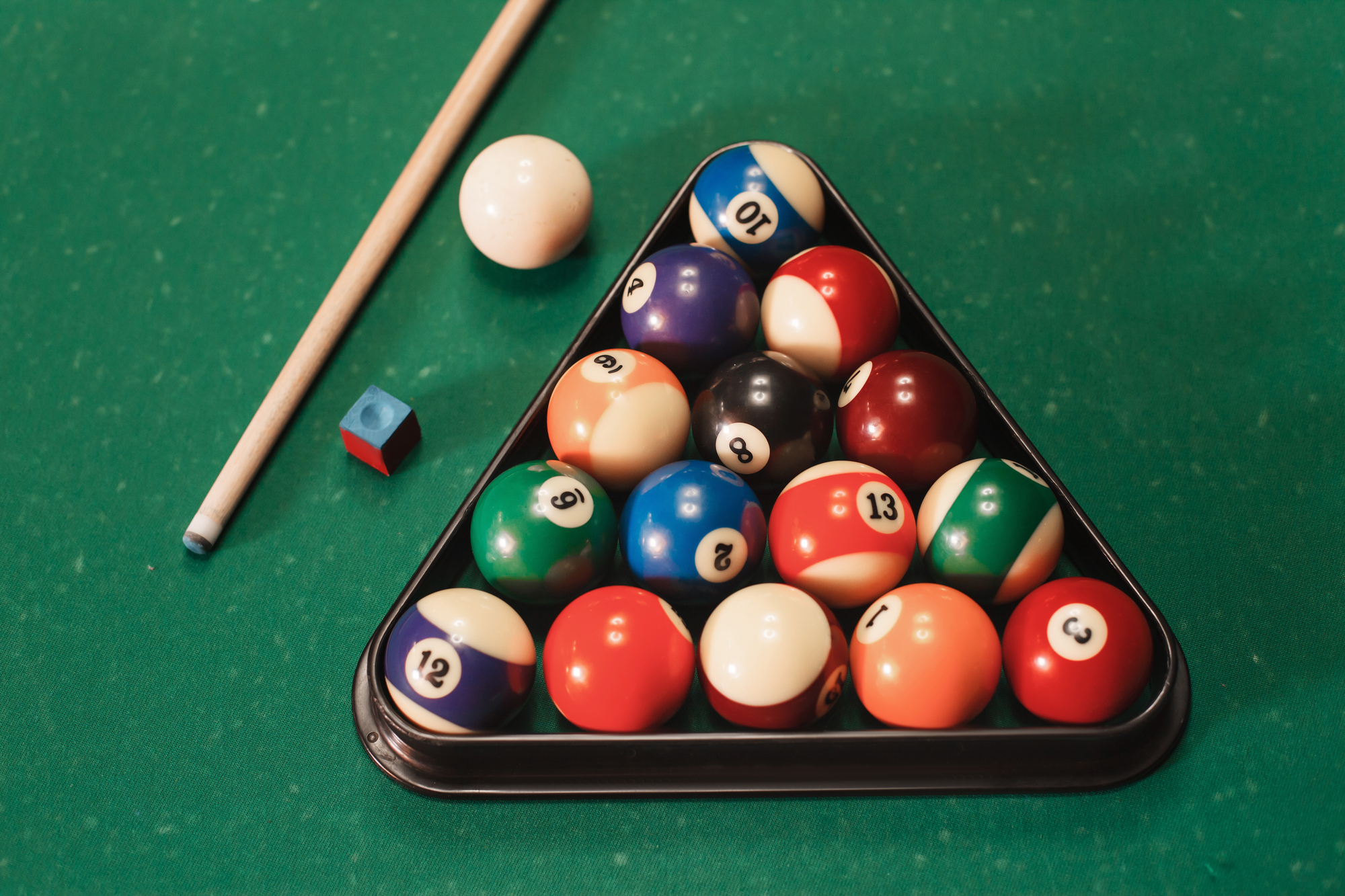 An image of pool table balls in a rack along with a cue ball, cue stick, and chalk on a pool table